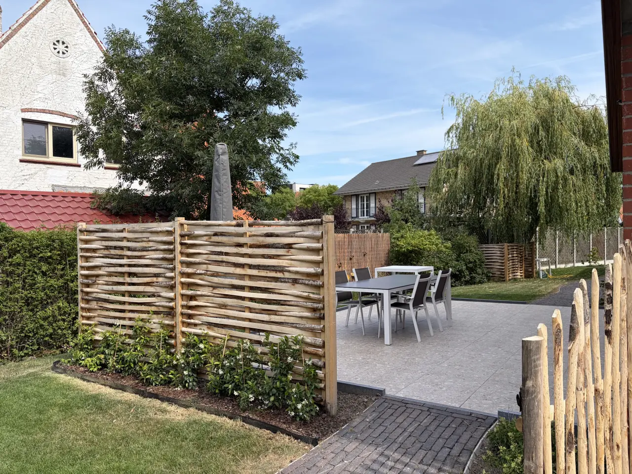 Een buitentafel met stoelen op het afgesloten terras in de tuin, met een gevlochten houten scherm en groen op de achtergrond.